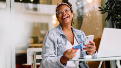 Mulher negra sorrindo com o celular e cartão na mão frente ao notebook