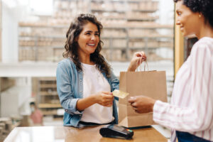 Mulher dando o cartão para vendedora com sacola de compras na mão