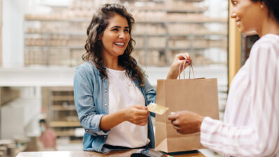 Mulher dando o cartão para vendedora com sacola de compras na mão