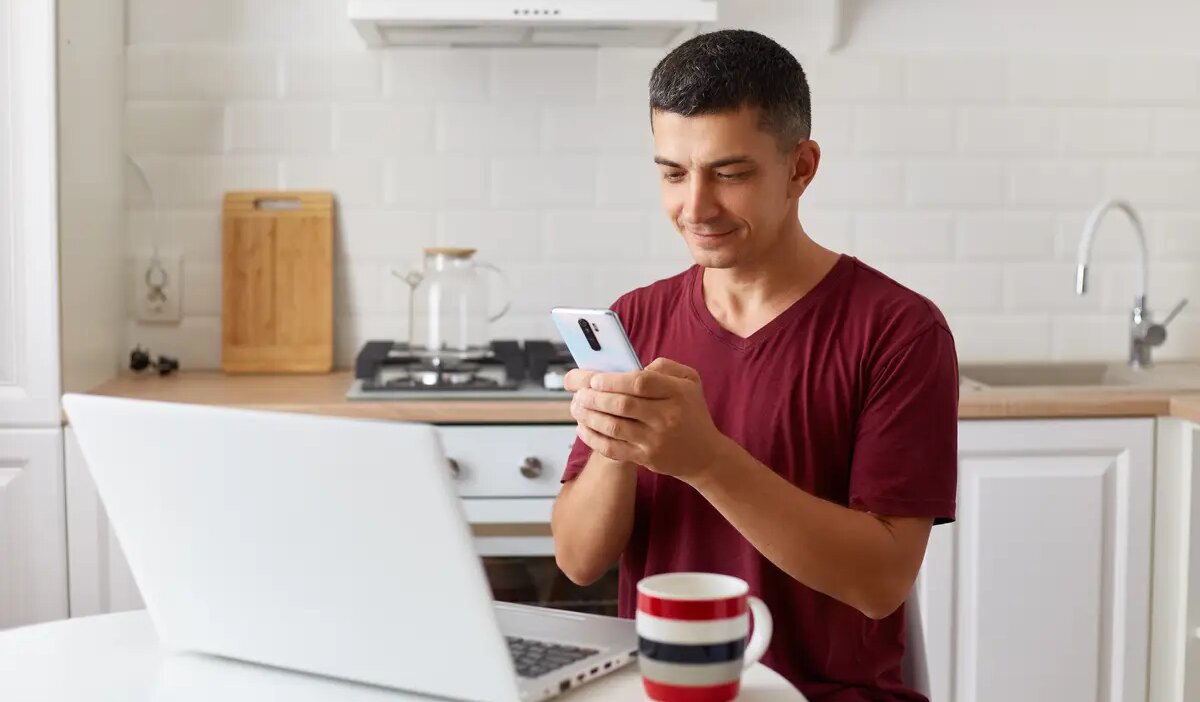 Positive handsome man working online at home on laptop, using smartphone for checking e-mails while having break fro freelance work, looking at device screen, typing message.