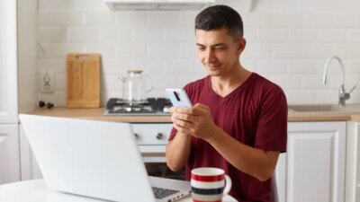 Positive handsome man working online at home on laptop, using smartphone for checking e-mails while having break fro freelance work, looking at device screen, typing message.